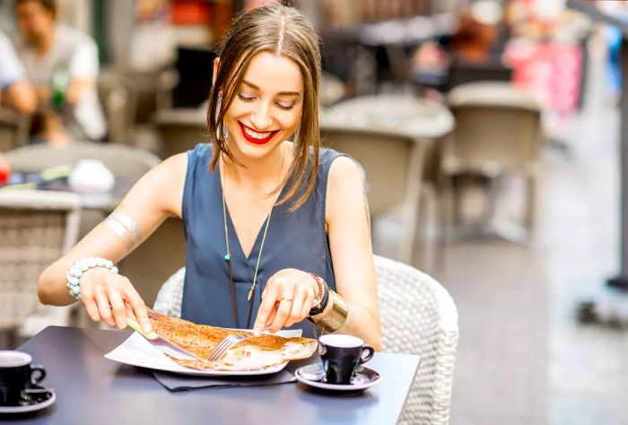 A woman beams as she cuts into her crêpe at an outdoor café.