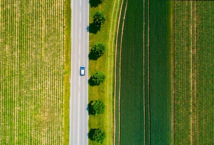 A grey vehicle drives along a concrete road bordered by trees amidst expansive green fields.