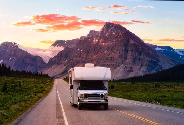 A white camper van cruising down an empty road at sunrise, surrounded by snow-capped mountains.