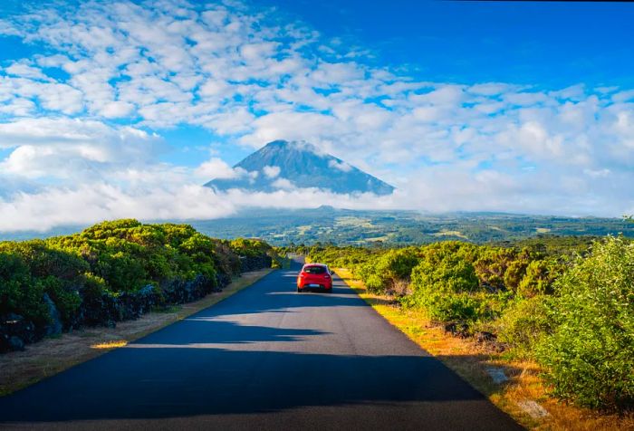 A red car drives along a road that stretches across a lush landscape, with distant mountains shrouded in clouds.