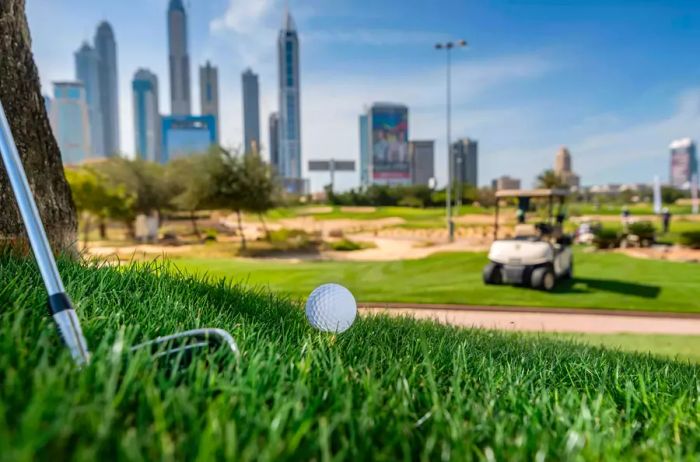 A day on the golf course. Close-up of a golf ball resting on lush green grass, with Dubai's skyscrapers in the background alongside a golf cart.