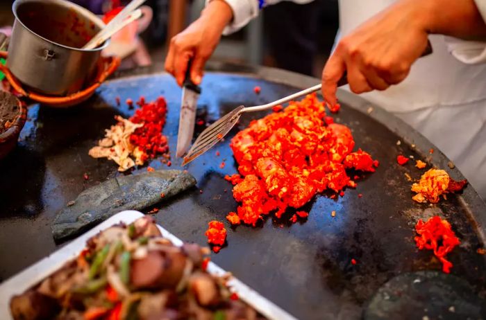 A quesadilla is being prepared on a large comal, which resembles a flat iron heated over fire, akin to a barbecue grill. Chorizo is also sizzling, likely to be added to the dish.