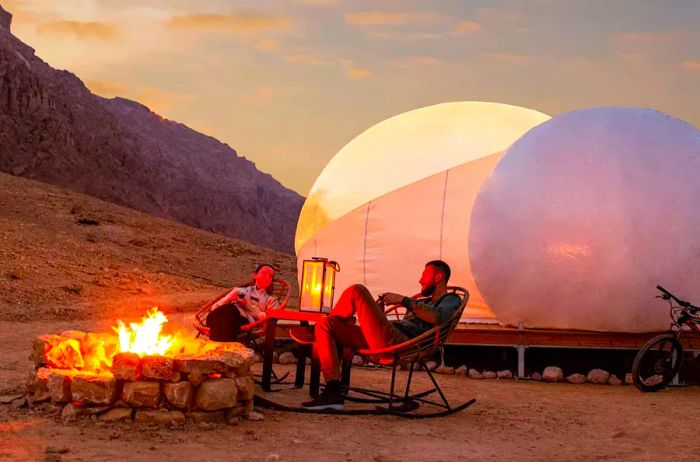 Two individuals gather around a campfire near dome-shaped tents in Jebel Hafit Desert Park.