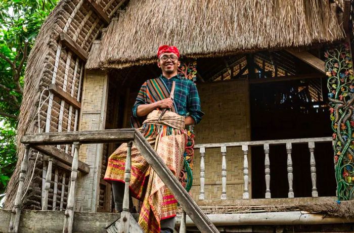 A young male tourist dressed in traditional Lombok attire posing in front of a traditional Sasak house.