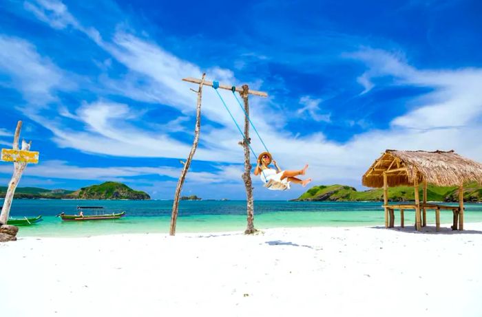 A woman in a white dress and hat enjoying a swing at a tropical beach under sunny skies.