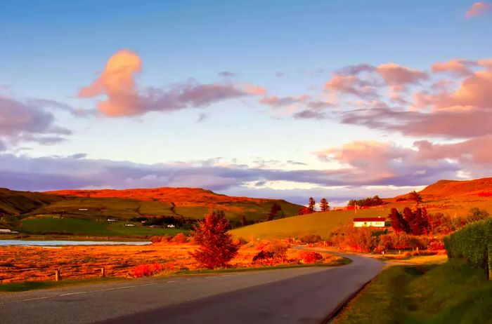 The golden hour before sunset bathed the green hills, dotted with homes along a winding road on the Isle of Skye in Scotland, in a warm orange glow.