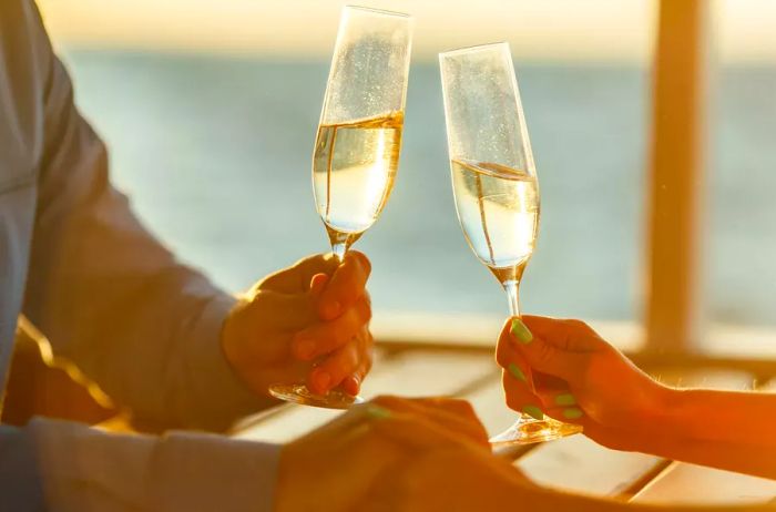 A couple joyfully clinks champagne glasses with one hand while holding each other's hands with the other.