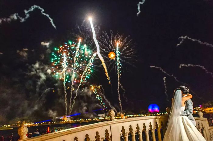 A heterosexual couple celebrating their wedding day at Disney World with a backdrop of fireworks lighting up the sky