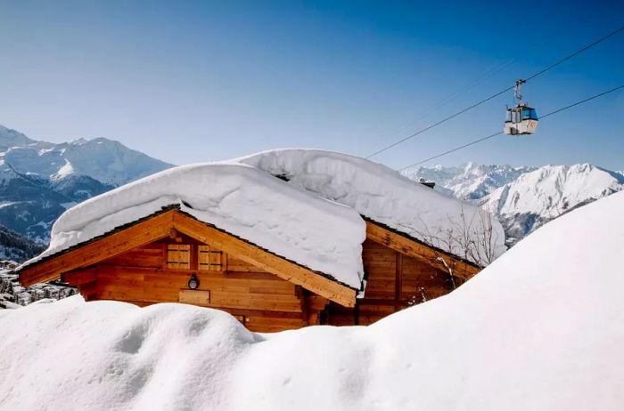 A picturesque, snow-laden wooden chalet basks in the sunlight, with a ski lift in the background and stunning blue Swiss mountains blanketed in snow.