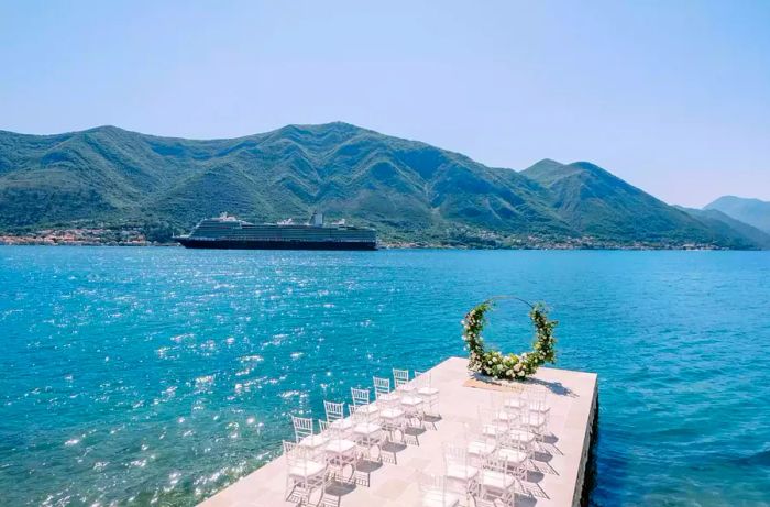 A row of white chairs is arranged in front of a circular wedding arch by the sea, with a cruise ship passing in the background.