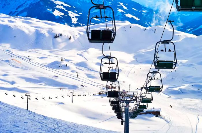 A sizable ski chairlift ascends a steep slope marked by tracks on the snowy peak of a high mountain in the Austrian Alps, alongside a quaint chalet.