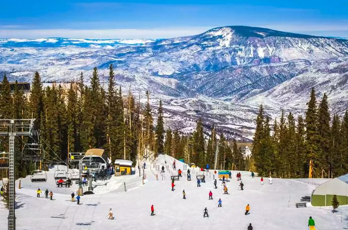 A clear winter day reveals a stunning view of a Colorado ski slope, where people are skiing and snowboarding down to the base of the chairlift, surrounded by a backdrop of forests and mountains.