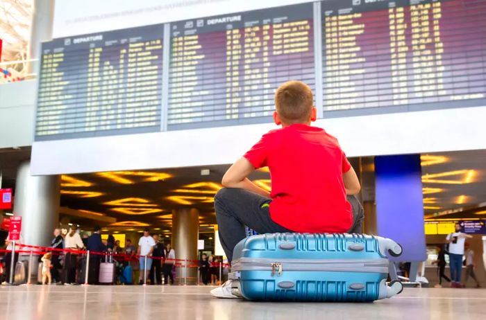 A lonely child sitting on a suitcase, waiting for departure at the airport while looking at the arrivals board.