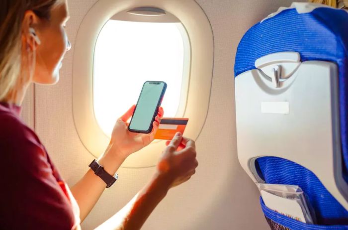 A young woman seated by the airplane window during a flight, holding a credit card and making an online payment through her smartphone.