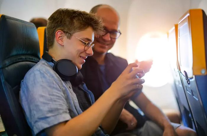 A father and his teenage son traveling by plane, with the boy showing his father a game on his smartphone.