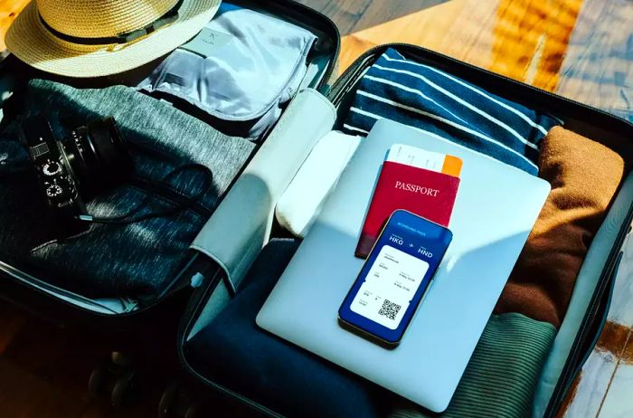 A high-angle view of an open suitcase filled with clothes, a sun hat, a camera, a laptop, and a passport on a wooden floor illuminated by sunlight, alongside a smartphone displaying an electronic flight ticket itinerary. Travel essentials. Travel and vacation theme.