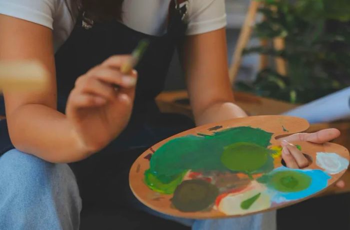 Close-up of a female artist at an easel, dipping her brush into a colorful palette.