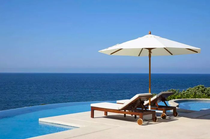 Lounge chairs shaded by an umbrella beside an infinity pool with ocean views