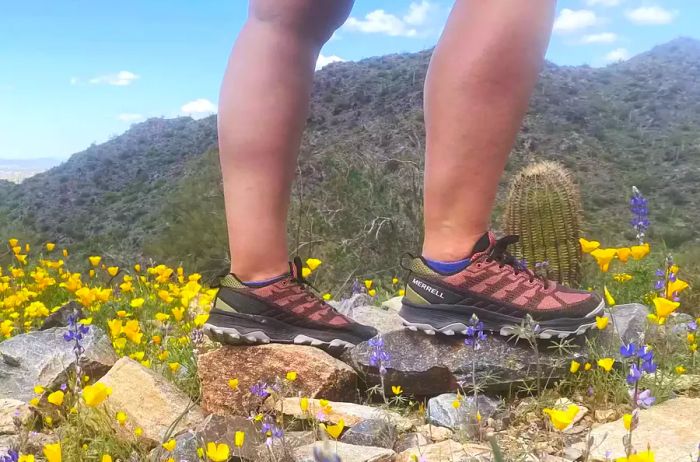 A side view of a woman standing on rocks, wearing Merrell Speed Eco Hiking Shoes, surrounded by vibrant yellow and purple flowers.