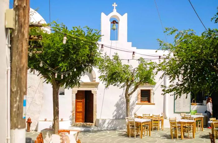 Empty white restaurant tables set in front of an old Greek Orthodox church nestled in a backstreet of Ano Meria town.