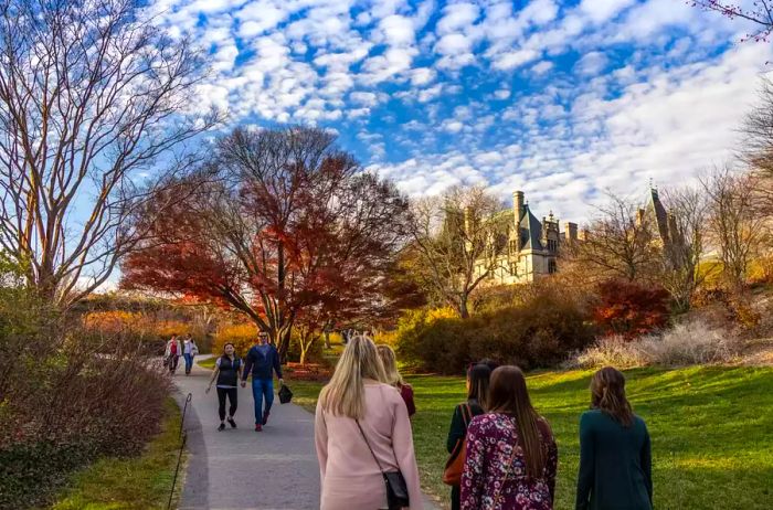 Visitors strolling along a path with the Biltmore Estate in the background, framed by trees. Captured during the Christmas at Biltmore celebration, though no holiday decor is visible. Some autumn colors and bare winter trees are present.