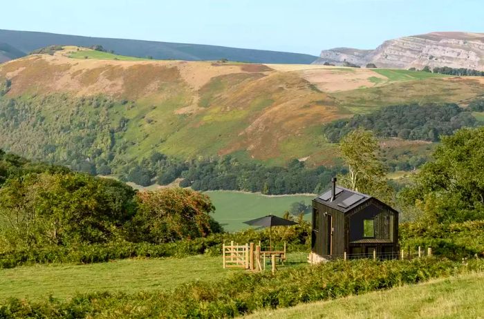 A sleek black cabin is nestled in the Welsh countryside, offering views of a nearby hill