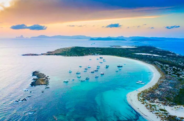 Aerial view of Espalmador and Ibiza at sunset, featuring sailboats anchored in the bay.