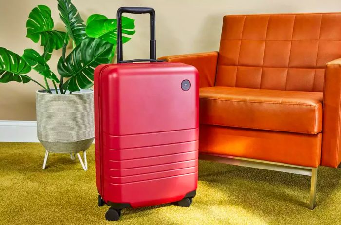 Monos Carry-On Plus positioned beside a chair and plant on a green carpet