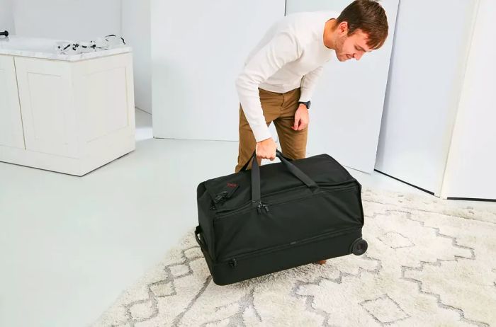 A person lifts the Tumi Alpha 3 Large Split 2-Wheeled Duffel off a white and gray carpet