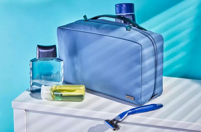 Away Large Toiletry Bag displayed on a dresser alongside cologne, a mini shampoo bottle, a razor, and lotion