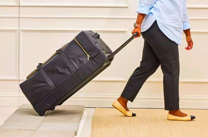 A person rolls the Mark & Graham Terminal 1 Checked Rolling Duffle Bag across a stone surface