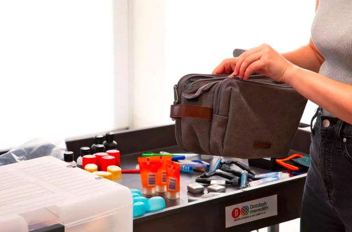 A hand is seen unzipping a Bagsmart Toiletry Bag for Men on a testing cart filled with mini travel-sized products.