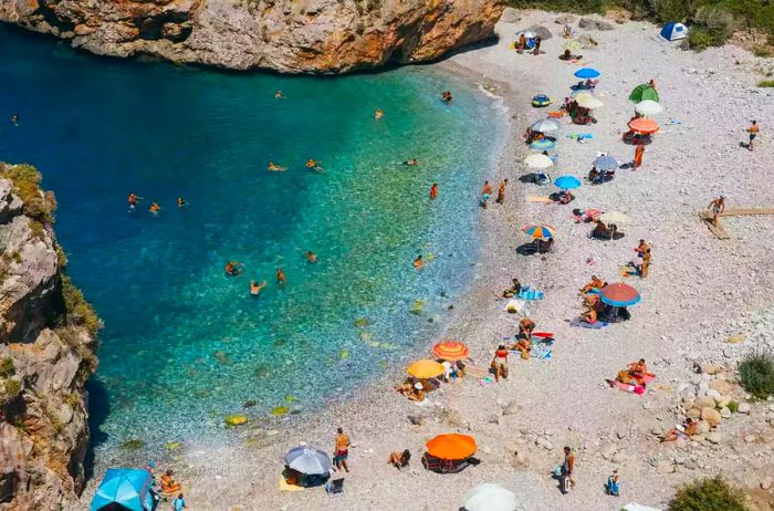 A quaint rocky beach in Kardamyli, Greece, filled with sunbathers, vibrant umbrellas, and shimmering turquoise water.