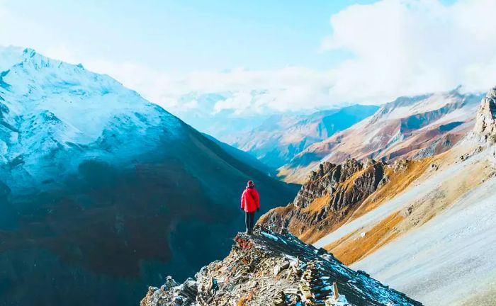 A person stands atop a mountain, gazing down at a steep drop into the valley below.