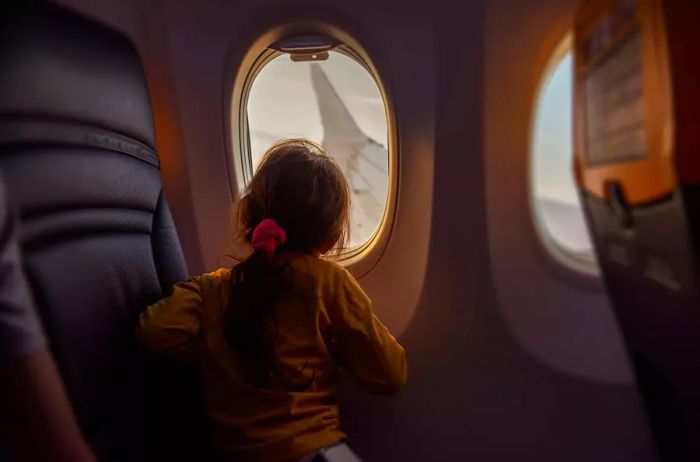 A young girl gazes out the airplane window at the wing. She feels fearless flying, peering at the sky through the porthole. This captures the curiosity and bravery children exhibit while flying. A glimpse of the airplane's interior atmosphere.