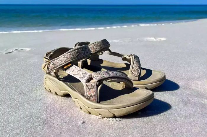 A pair of Teva Terra Fi 5 Universal Sandals rests on the sand with the ocean in the background