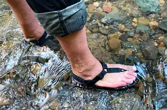 A person wades through a stream while wearing Bedrock Cairn Adventure Sandals