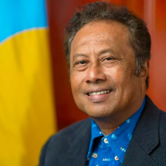 A portrait of a smiling Tommy Remengesau, Jr., president of Palau, posed in front of the Palau flag.