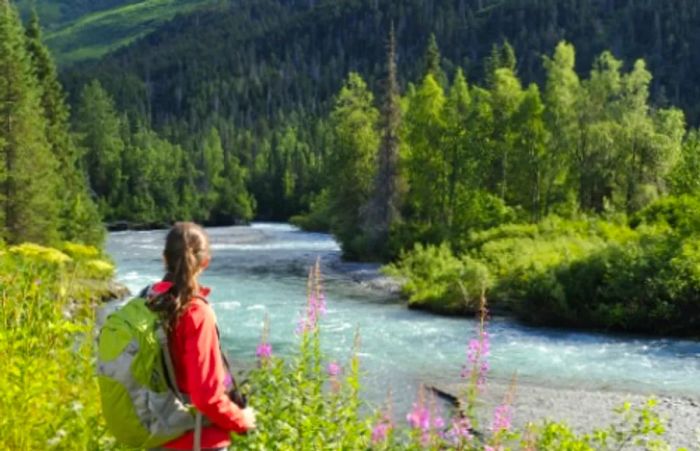 A woman in a red jacket hikes alongside a river in the Alaskan wilderness.