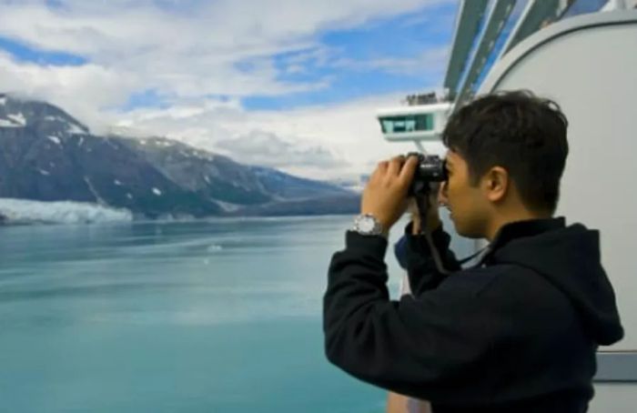 A person dressed in a black sweater gazes at the stunning Alaskan landscape through binoculars from a cruise ship balcony.