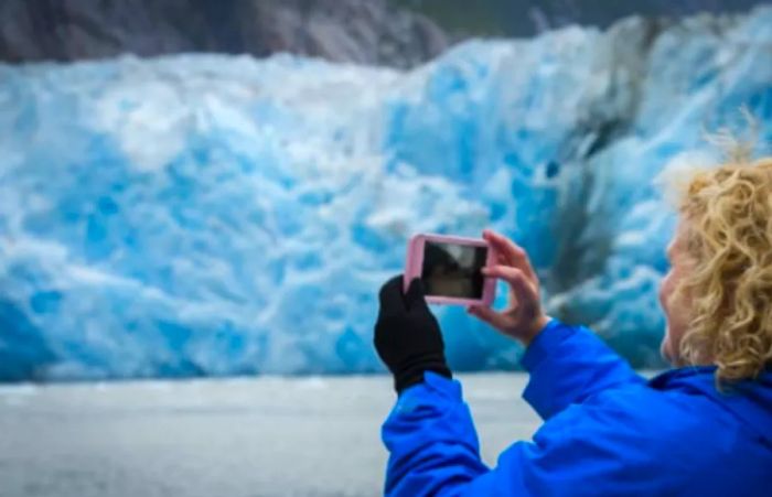 A woman in a blue jacket captures images of glaciers with her pink smartphone.