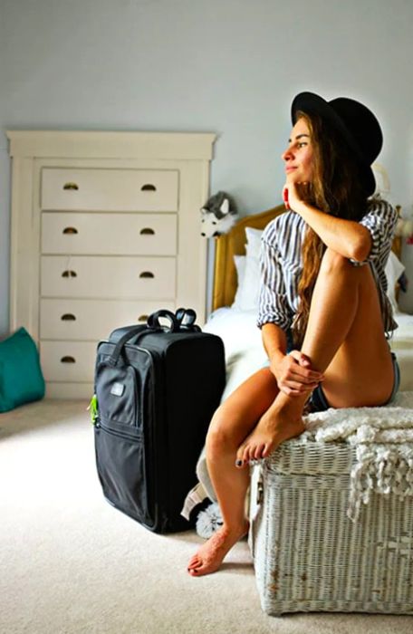 Andrea sitting on a bed, wearing a hat, with her suitcase nearby.