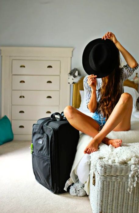 Andrea is seated on a bed, holding a hat in front of her face, with a suitcase beside her.