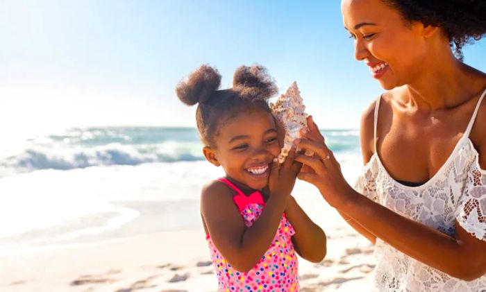 A joyful mother holding a seashell to her daughter's ear while enjoying a stunning beach.