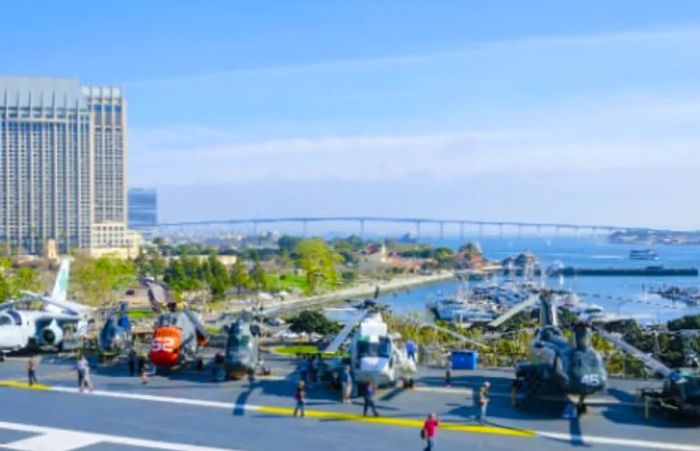 A tourist exploring various helicopters at the aircraft carrier museum in San Diego, with the Coronado Bridge in the background