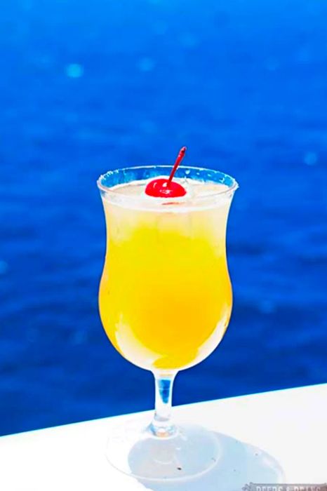 A vibrant orange drink topped with a red cherry, set against the backdrop of a bright blue ocean