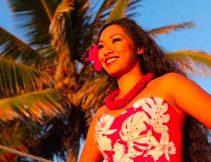 a cheerful young Hawaiian woman posing behind a palm tree with her hands on her hips