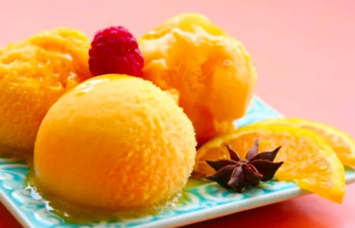 three scoops of sherbet arranged on a blue plate alongside orange slices and a raspberry