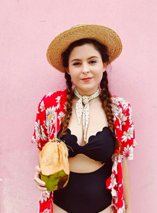 Noelle in a swimsuit sipping from a coconut against a vibrant pink wall