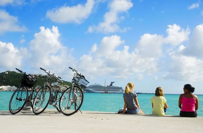 Three women pause their bike ride to admire the St. Maarten coastline, with a Dinogo cruise ship docked in the background.